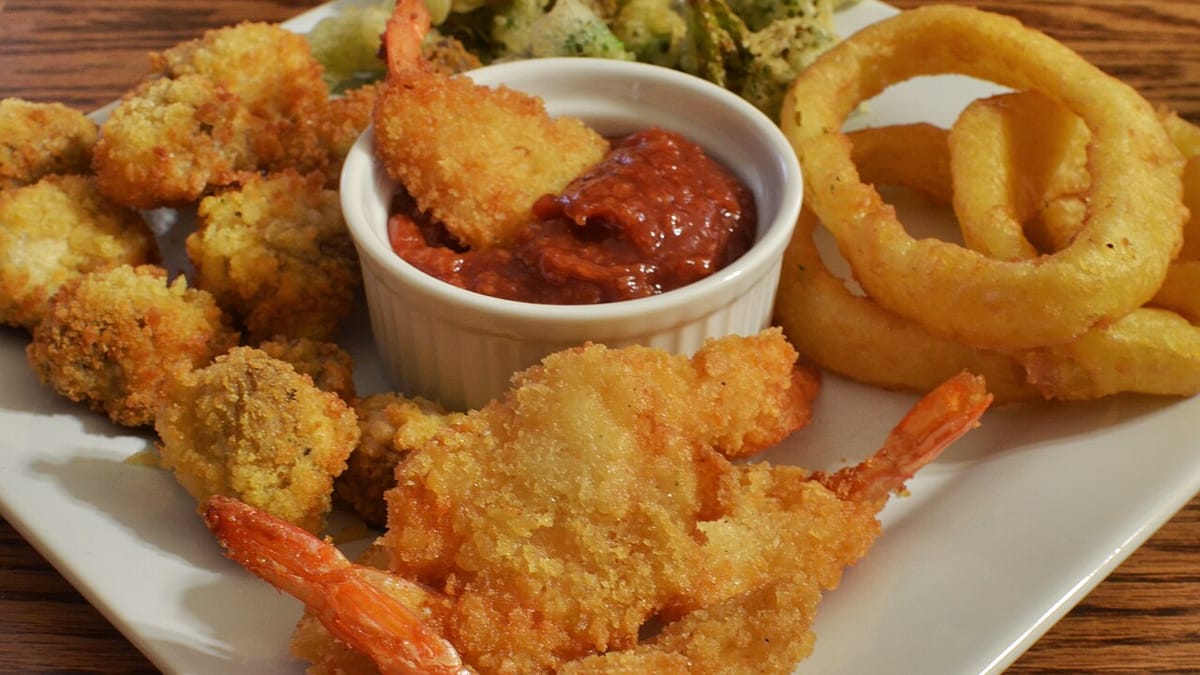 Sepiring gorengan berminyak berisi udang goreng tepung, onion ring, dan aneka gorengan dengan semangkuk saus merah