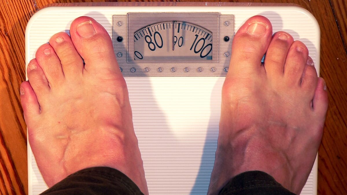 Sepasang kaki berdiri di atas timbangan badan analog yang jarumnya menunjukkan angka sekitar 90 kilogram.