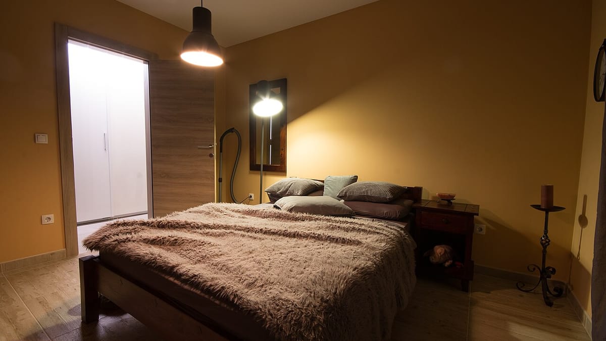 Kamar tidur sederhana bercahaya hangat dengan kasur berselimut bulu cokelat, beberapa bantal, lampu gantung, dan lampu berdiri