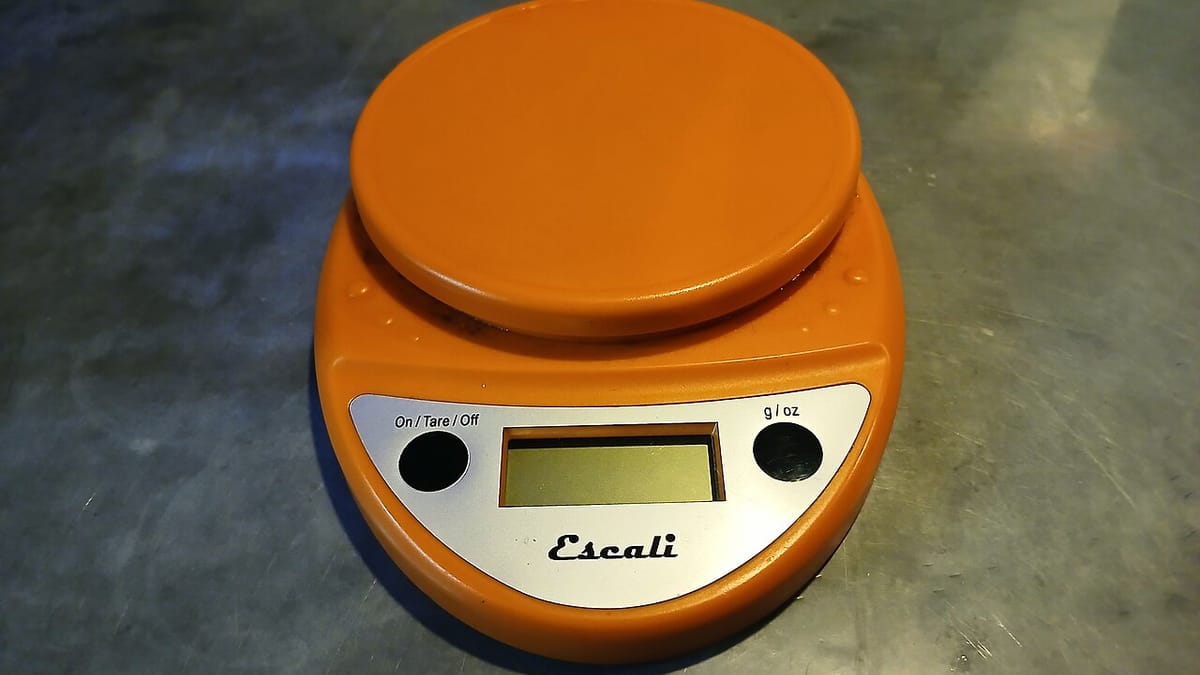 Timbangan dapur digital berwarna oranye dengan layar LCD di atas meja logam, alat bantu menimbang porsi makanan.
