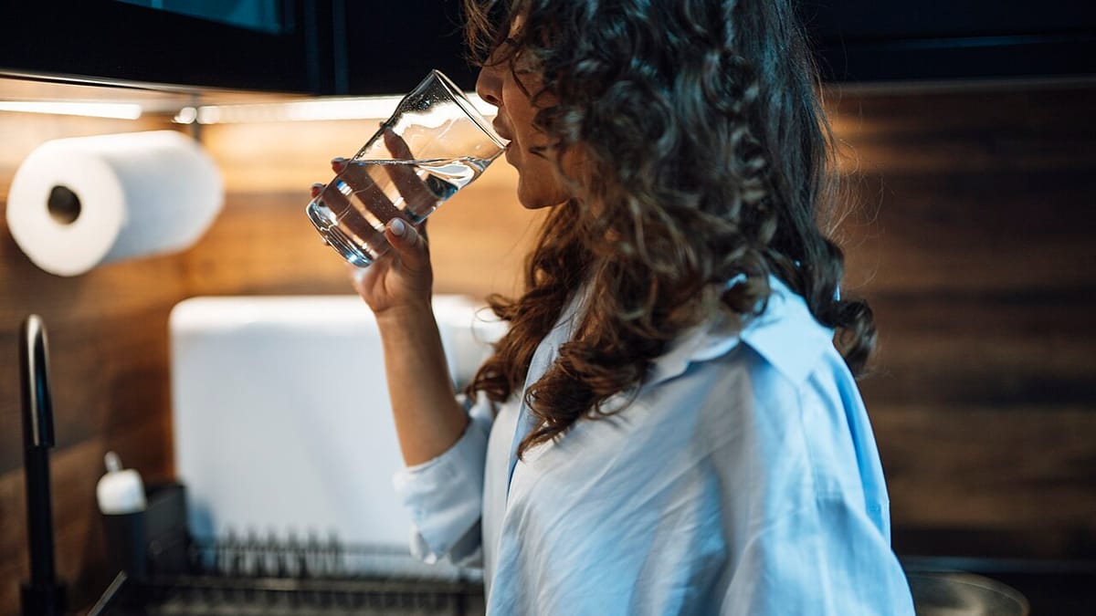 Perempuan berdiri di dapur sambil minum segelas air putih.