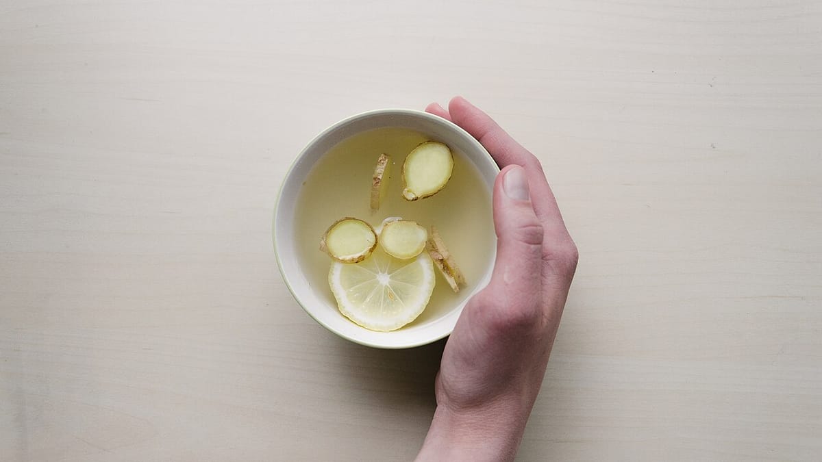 Tangan memegang cangkir putih berisi air hangat dengan irisan jahe dan lemon di atas meja berwarna terang.