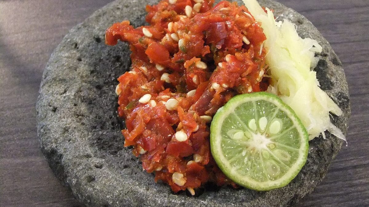 Sambal cabai merah segar bertabur biji cabai di atas cobek batu, dilengkapi irisan jeruk limau dan parutan buah.