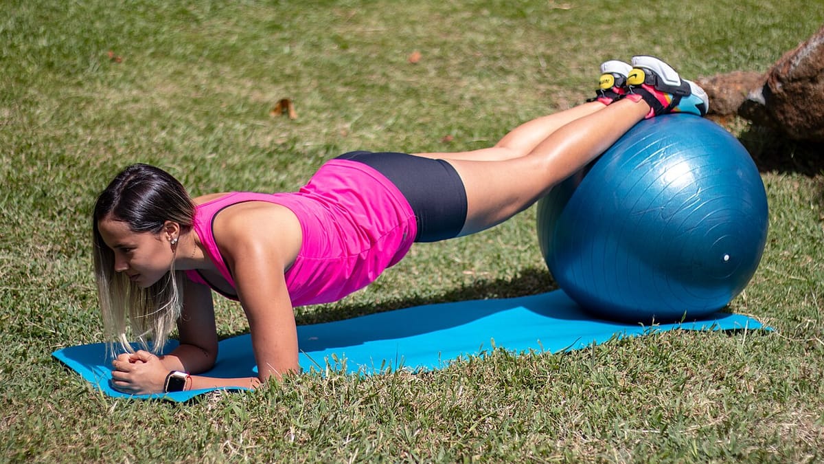 Seorang perempuan melakukan plank di atas matras biru di rumput dengan kedua kaki bertumpu pada gym ball berwarna biru