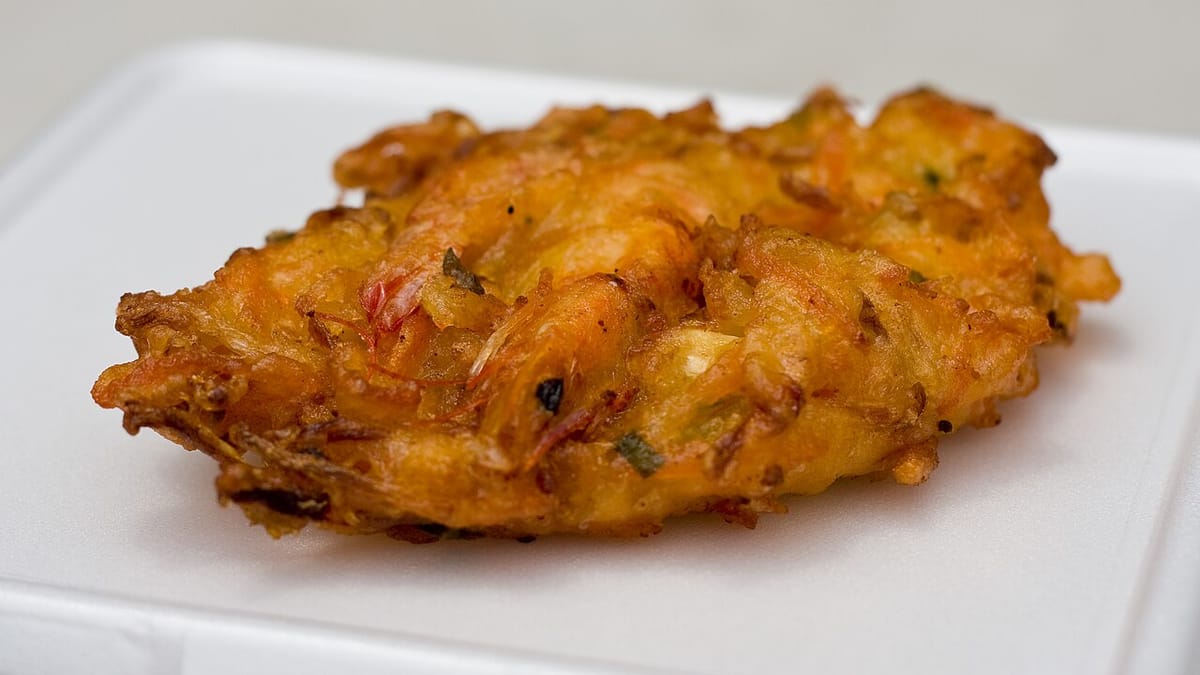 Sepotong bakwan goreng berwarna oranye kecokelatan berisi udang dan sayuran, disajikan di atas piring putih