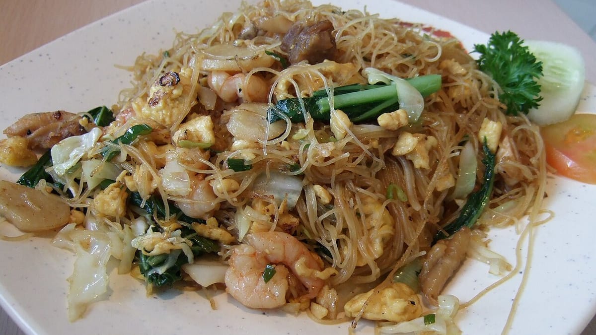 Sepiring bihun goreng dengan udang, telur orak-arik, kol, dan sawi hijau, dihiasi irisan timun, tomat, dan daun peterseli.
