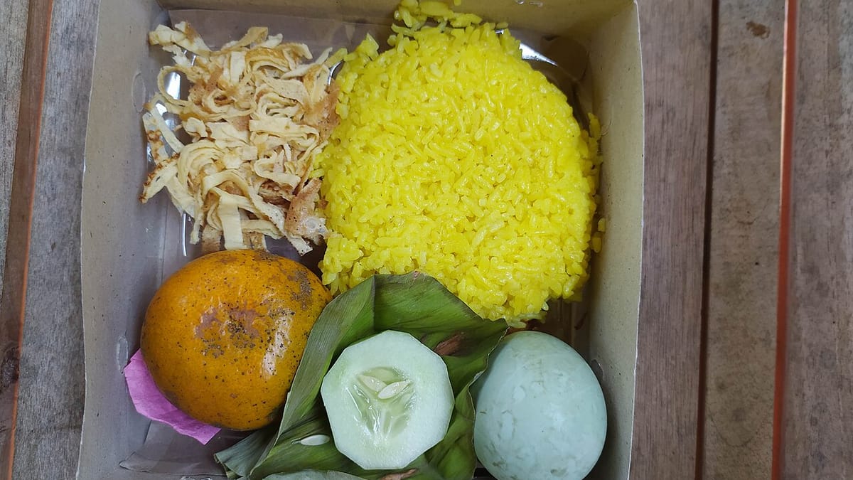 Nasi kuning dalam kotak kertas lengkap dengan suwiran telur dadar, telur rebus, irisan mentimun, dan sebuah jeruk.
