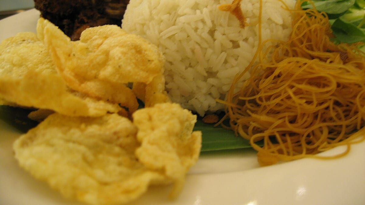 Sepiring nasi uduk bertabur bawang goreng disajikan dengan emping, bihun goreng, dan semur daging di atas daun pisang.