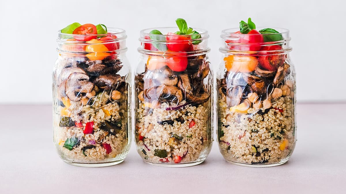 Tiga toples kaca berisi salad quinoa dengan jamur, kacang arab, tomat ceri, dan daun basil sebagai bekal porsi terukur.