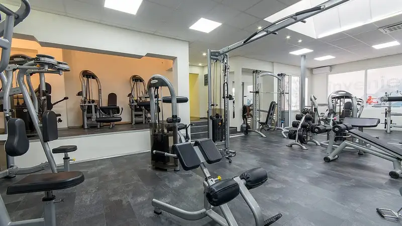 Interior gym modern berlantai abu-abu dengan deretan mesin beban, kabel crossover, dan bangku latihan tanpa pengunjung.