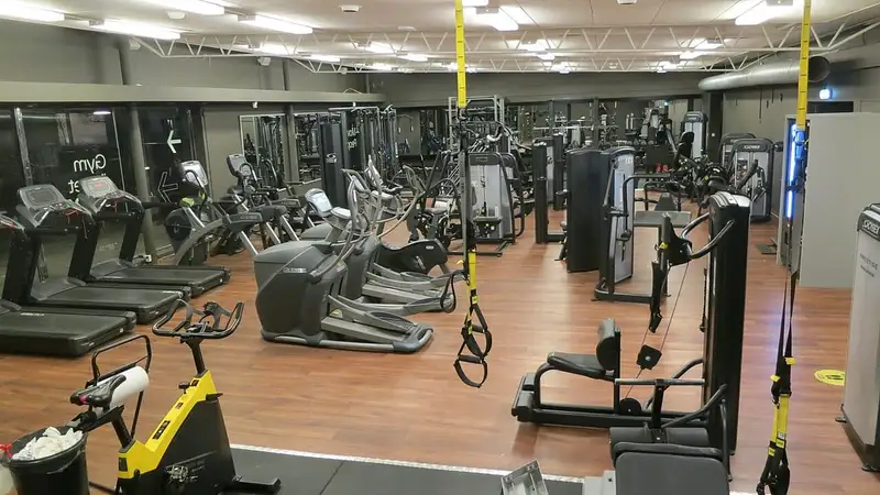 Ruang gym yang lengang berlantai kayu dengan deretan treadmill, sepeda statis, mesin beban, dan tali latihan suspensi.