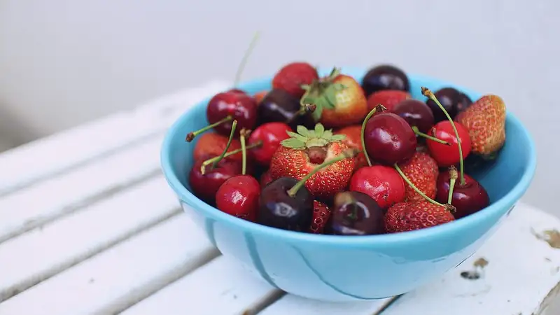 Semangkuk biru berisi buah ceri merah tua dan stroberi segar di atas bangku kayu putih