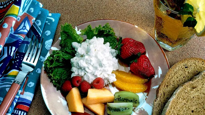 Sepiring keju cottage dengan stroberi, raspberry, jeruk, kiwi, dan melon di atas daun selada, ditemani roti gandum dan es teh.
