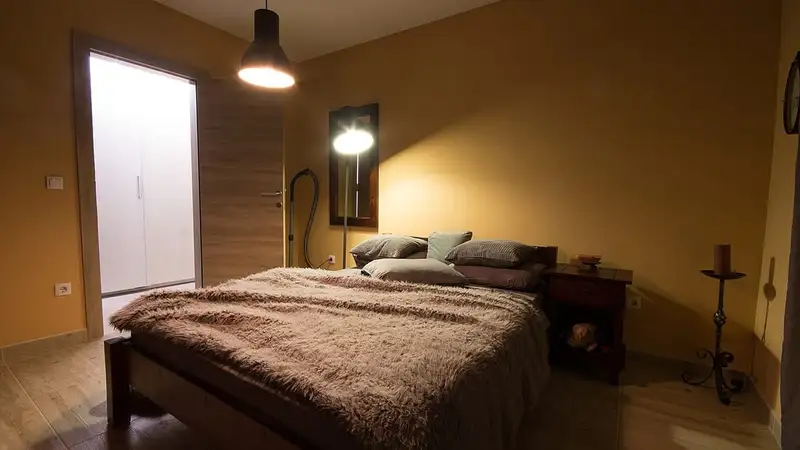 Kamar tidur sederhana bercahaya hangat dengan kasur berselimut bulu cokelat, beberapa bantal, lampu gantung, dan lampu berdiri