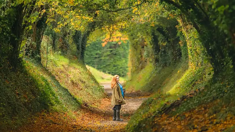 Seorang perempuan berjaket hijau dan bersepatu bot karet berdiri sambil menoleh ke belakang di jalan setapak berlapis daun gugur, di dalam lorong pepohonan rimbun di Halnaker, West Sussex, Inggris.