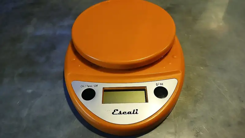 Timbangan dapur digital berwarna oranye dengan layar LCD di atas meja logam, alat bantu menimbang porsi makanan.