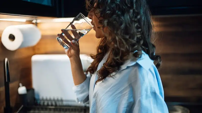 Perempuan berdiri di dapur sambil minum segelas air putih.