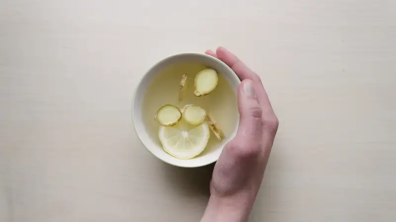 Tangan memegang cangkir putih berisi air hangat dengan irisan jahe dan lemon di atas meja berwarna terang.