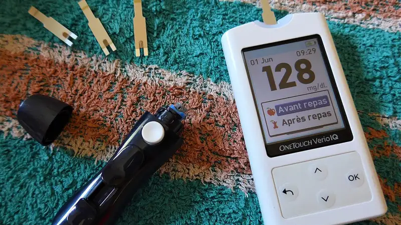 Alat pengukur gula darah (glukometer) menampilkan angka 128 mg/dL, berdampingan dengan pena lanset dan beberapa strip tes.