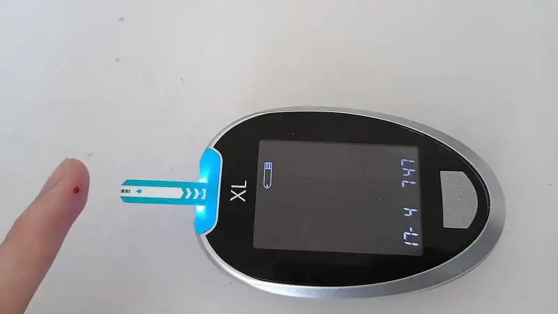 Glukometer digital hitam dengan strip tes yang menyala biru, di samping ujung jari dengan setetes kecil darah untuk cek gula darah.