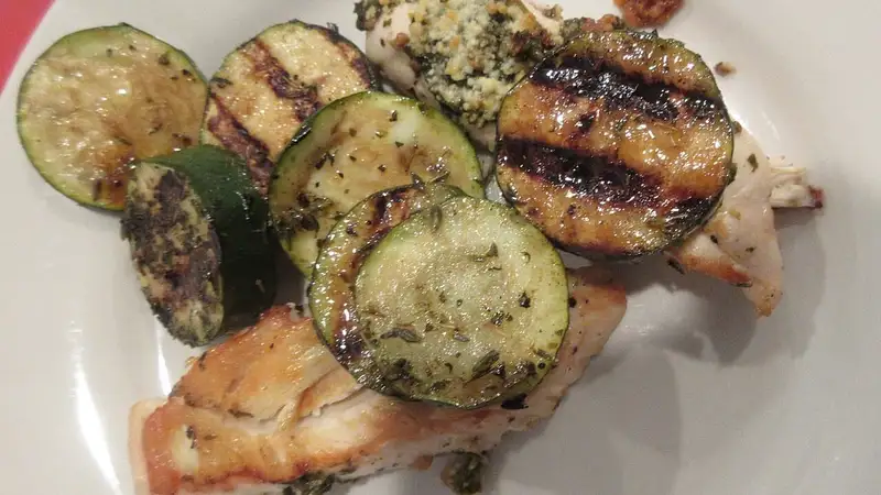 Dada ayam panggang berbumbu pesto bersama irisan zukini bakar bergaris panggangan di atas piring