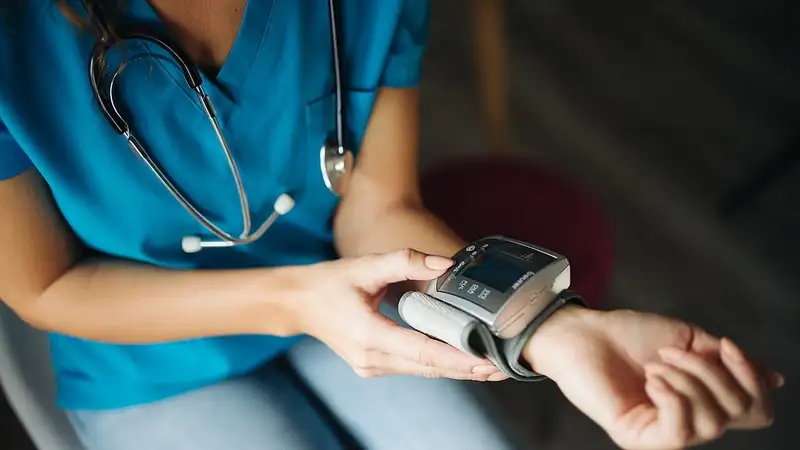 Tenaga kesehatan berseragam biru dengan stetoskop di leher mengukur tekanan darah di pergelangan tangannya dengan alat digital.