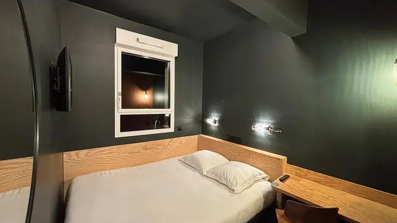 Kamar tidur hotel yang temaram dengan kasur putih, dua bantal, lampu baca dinding menyala, dan jendela kecil di dinding gelap.