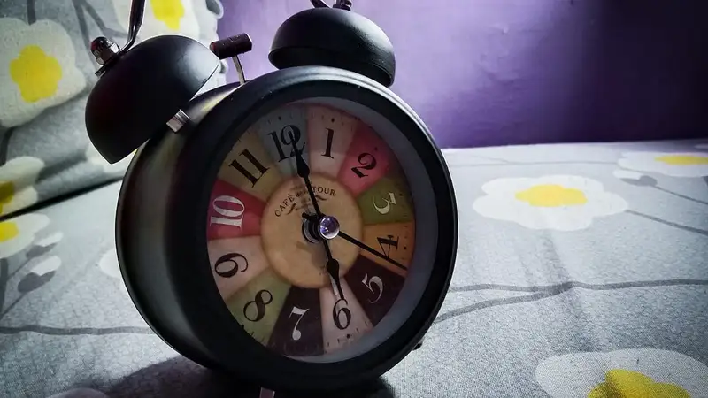 Jam weker hitam dengan dua lonceng dan angka berwarna-warni berdiri di atas seprai bermotif bunga di kasur