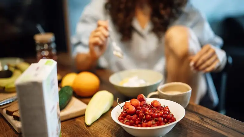 Seorang perempuan sarapan di meja kayu dengan semangkuk ceri dan kismis merah, yogurt, jeruk, timun, dan secangkir kopi