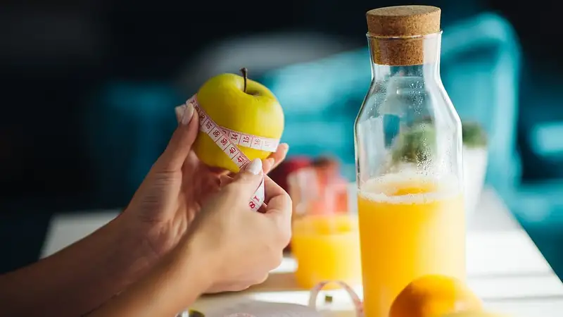 Tangan melilitkan pita ukur pada apel hijau di samping botol kaca dan gelas berisi jus jeruk segar di dapur yang terang.