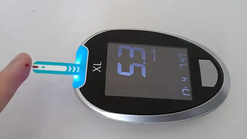 Alat pengukur gula darah digital dengan strip tes terpasang di dekat ujung jari bertetes darah kecil, layarnya menampilkan 5,3 mmol/L.
