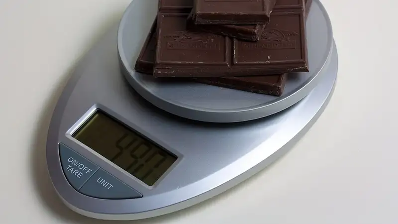 Potongan cokelat batangan ditimbang di atas timbangan dapur digital berwarna perak dengan layar angka dan tombol tare