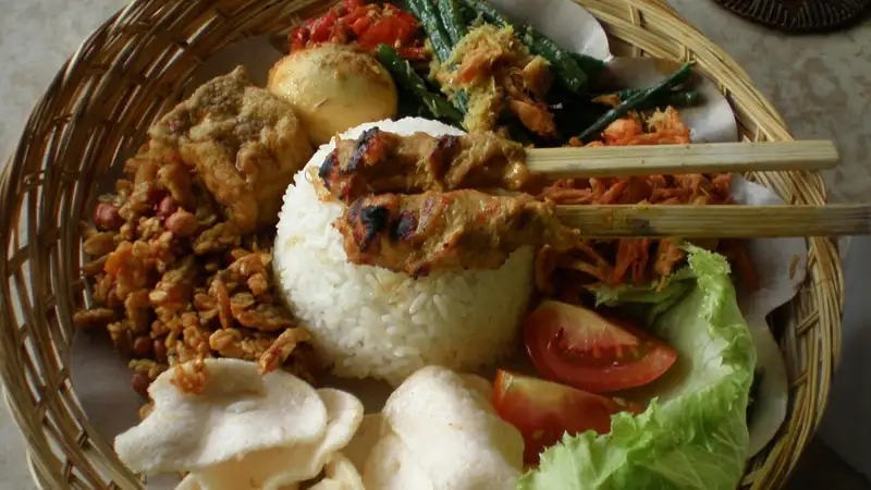 Nasi campur dalam wadah anyaman berisi nasi putih, dua tusuk sate, telur, kering tempe, buncis, tomat, selada, dan kerupuk.