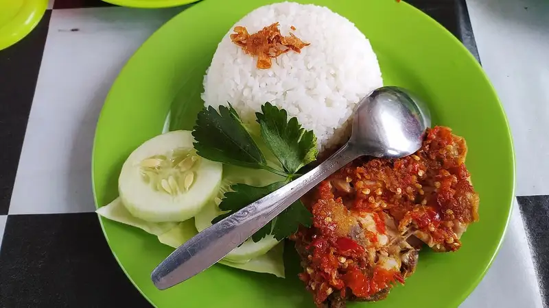 Sepiring nasi putih bertabur bawang goreng dengan ayam geprek berlumur sambal merah, irisan mentimun, dan daun seledri di piring hijau.