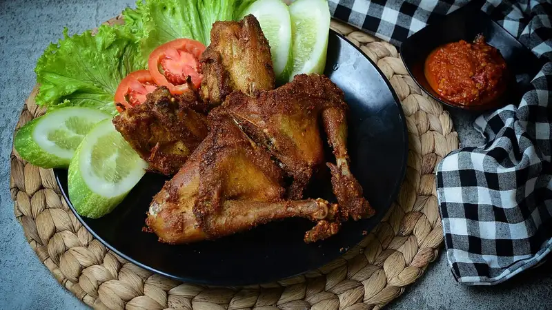 Potongan ayam goreng kalasan berwarna cokelat keemasan di piring hitam, disajikan dengan selada, timun, tomat, dan sambal.