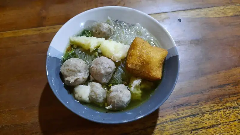 Semangkuk bakso berkuah berisi bola daging, bihun, tahu goreng, dan sayuran hijau di atas meja kayu.