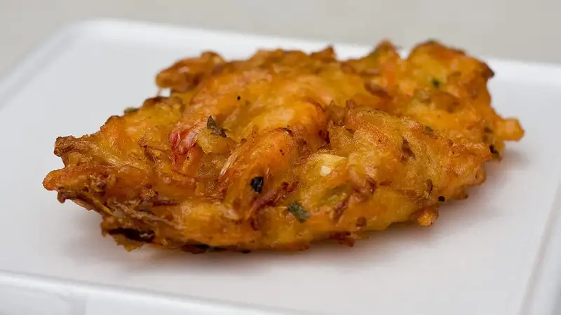Sepotong bakwan goreng berwarna oranye kecokelatan berisi udang dan sayuran, disajikan di atas piring putih