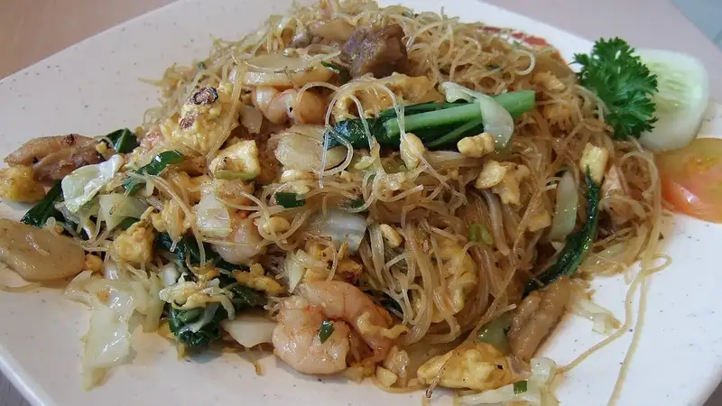 Sepiring bihun goreng dengan udang, telur orak-arik, kol, dan sawi hijau, dihiasi irisan timun, tomat, dan daun peterseli.