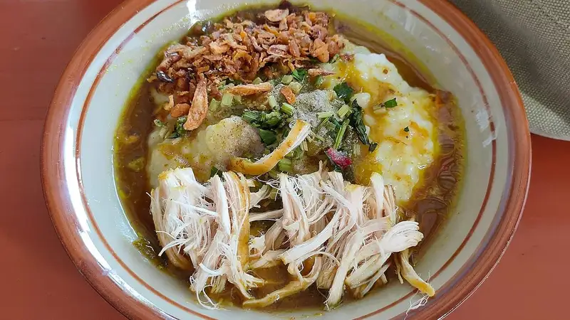 Semangkuk bubur ayam berkuah kuning dengan suwiran ayam, bawang goreng, irisan daun bawang, dan seledri di atas meja.