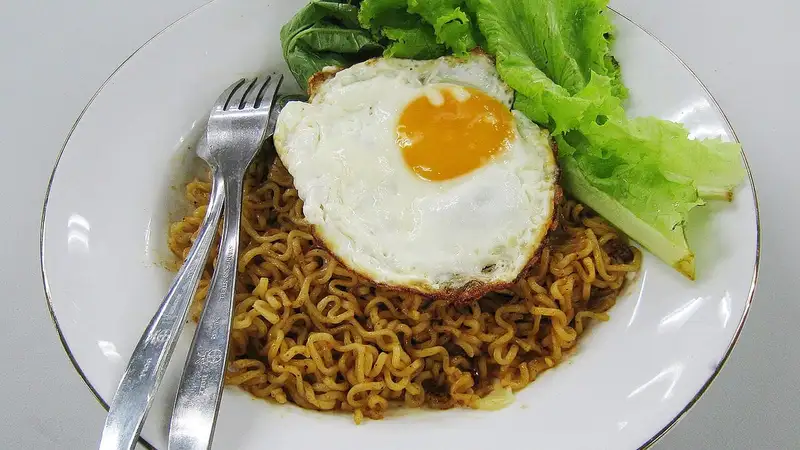 Sepiring mi goreng instan dengan telur mata sapi dan selada di atasnya, disajikan bersama garpu di piring putih