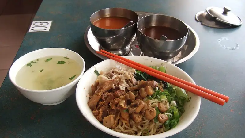 Semangkuk mie ayam dengan potongan ayam kecap dan daun bawang, disertai mangkuk kuah kaldu, sumpit merah, dan wadah saus