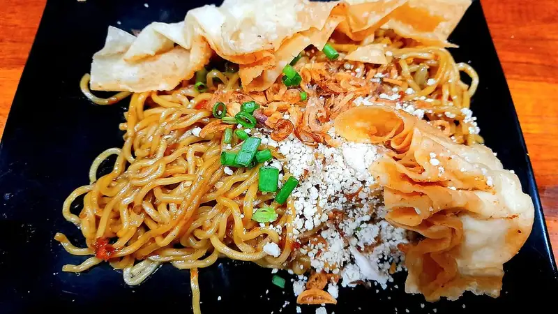 Seporsi mie pedas Mie Gacoan di atas piring hitam, ditaburi daun bawang dan dilengkapi pangsit goreng renyah.