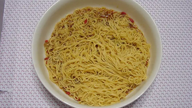 Semangkuk mi instan yang sudah dimasak dan tercampur bumbu kecokelatan, tersaji di mangkuk putih di atas taplak bermotif.