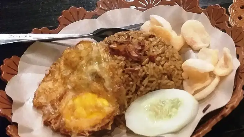Sepiring nasi goreng kecokelatan dengan telur ceplok, irisan mentimun, dan kerupuk putih di atas alas kertas.