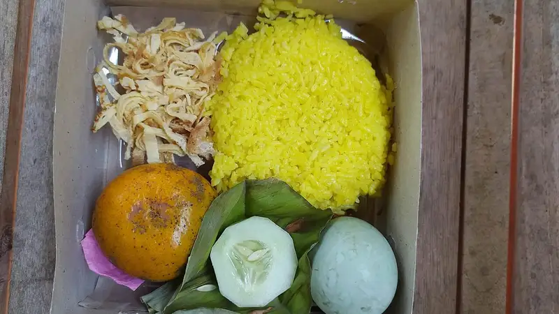 Nasi kuning dalam kotak kertas lengkap dengan suwiran telur dadar, telur rebus, irisan mentimun, dan sebuah jeruk.