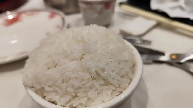 Semangkuk nasi putih pulen yang membukit di mangkuk putih, di atas meja makan restoran dengan sumpit dan peralatan makan di belakangnya.