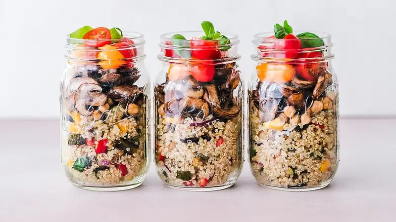 Tiga toples kaca berisi salad quinoa dengan jamur, kacang arab, tomat ceri, dan daun basil sebagai bekal porsi terukur.