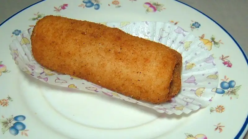 Sepotong risoles goreng berlapis tepung panir keemasan di atas kertas alas kue, disajikan di piring bermotif bunga.