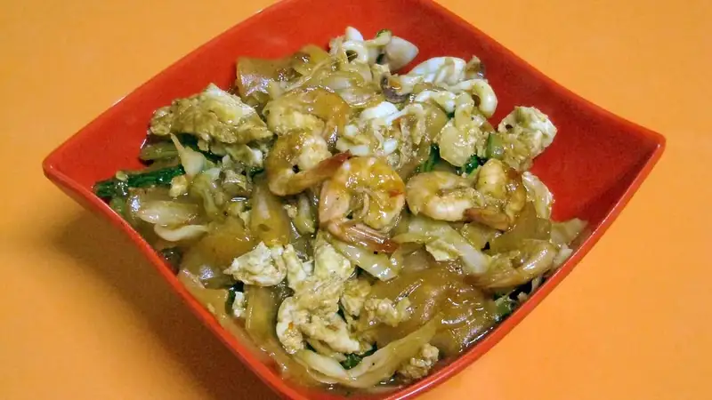 Seblak seafood berisi kerupuk basah, udang, dan telur orak-arik berbumbu dalam mangkuk merah di atas meja oranye