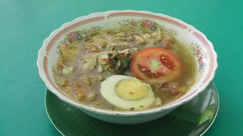 Semangkuk soto ayam berkuah bening berisi potongan ayam, setengah telur rebus, dan irisan tomat di atas piring alas hijau
